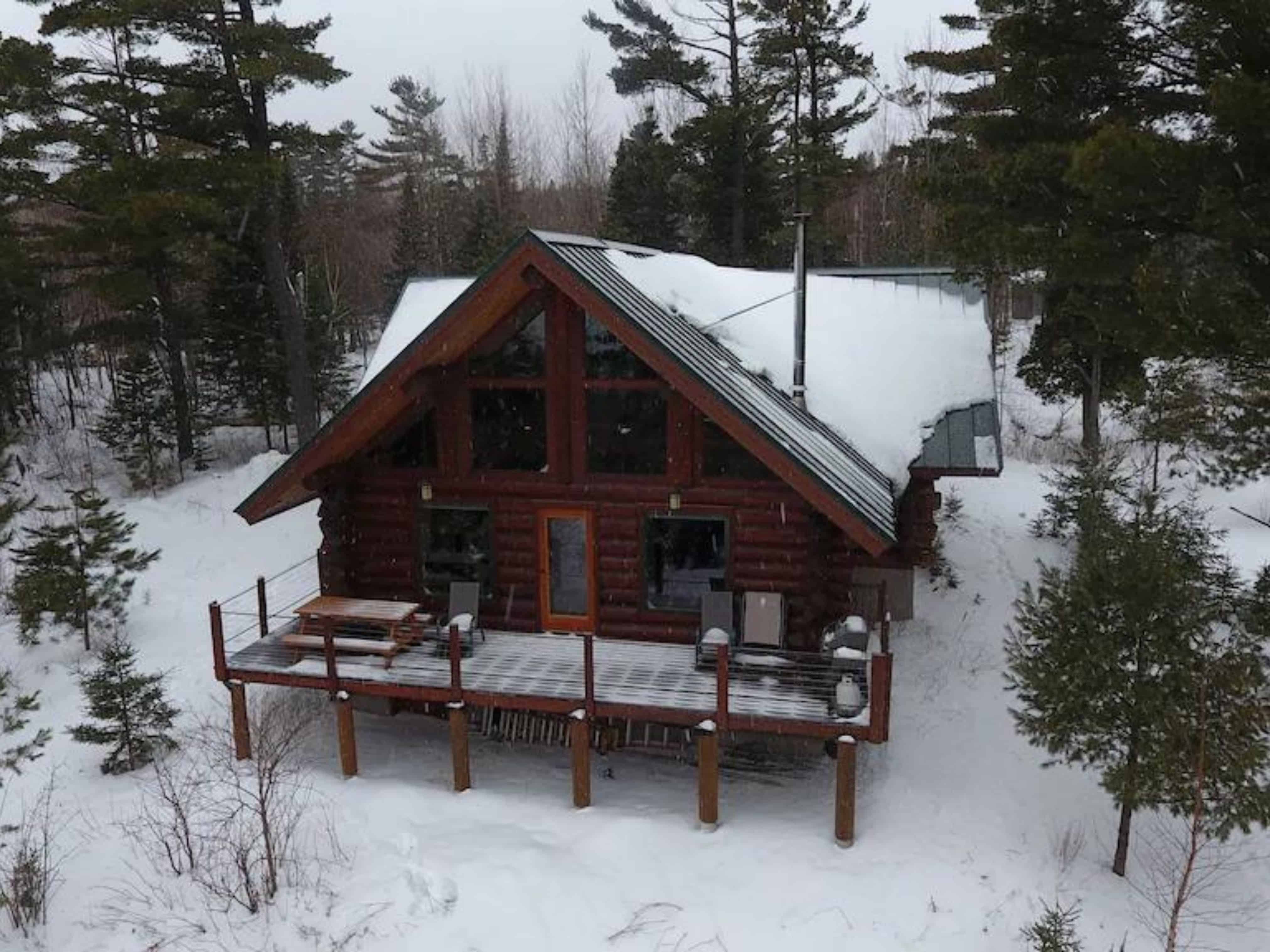 A large log cabin during winter with a front patio tucked away in the middle of the forest.